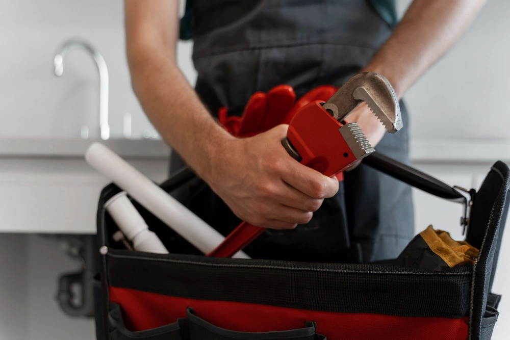 Plumber holding a red wrench over a tool bag with pipes in a kitchen setting.