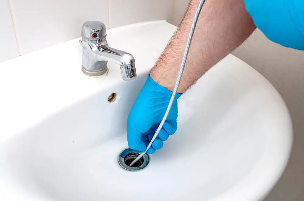 %Title% | Plumbing Champions A person wearing blue gloves uses a plumbing snake to unclog a bathroom sink drain next to a chrome faucet.
