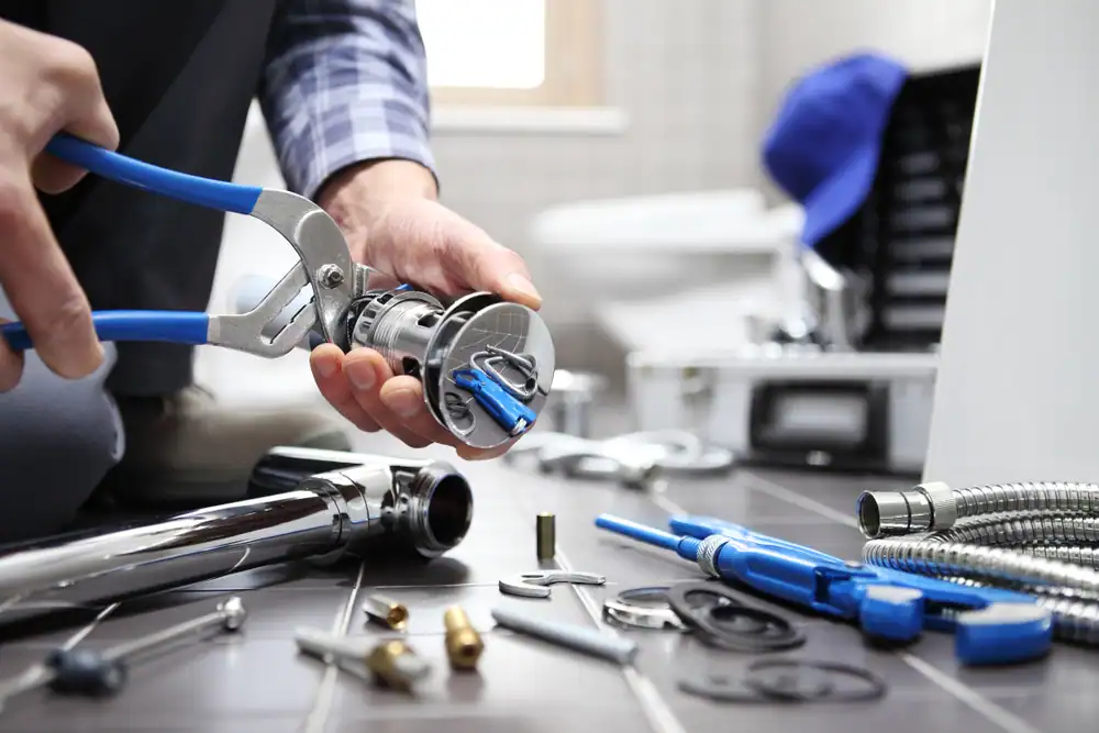 A person uses pliers to assemble plumbing parts on a tiled bathroom floor, surrounded by various tools, hoses, and fittings.