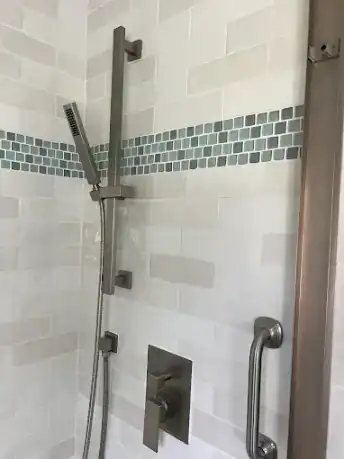 Shower wall with white subway tiles, a grey glass mosaic accent strip, and chrome fixtures including a slide bar showerhead and grab bar, shown during a bathroom remodel installation.
